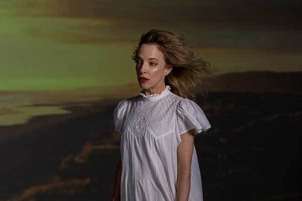 A woman in white nightgown stands on a cliff's edge as the sun sets.